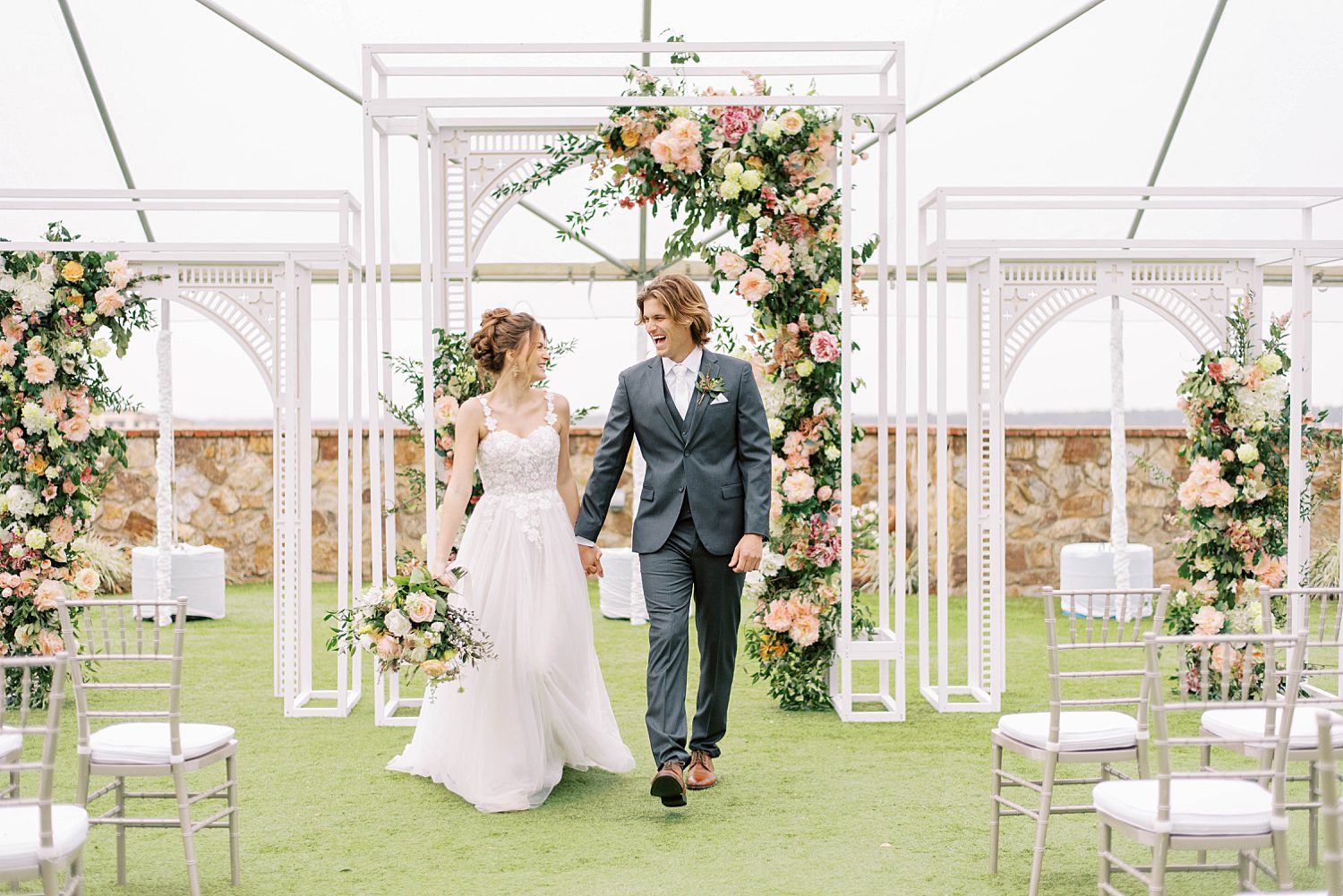 newlyweds hold hands walking through white arbor