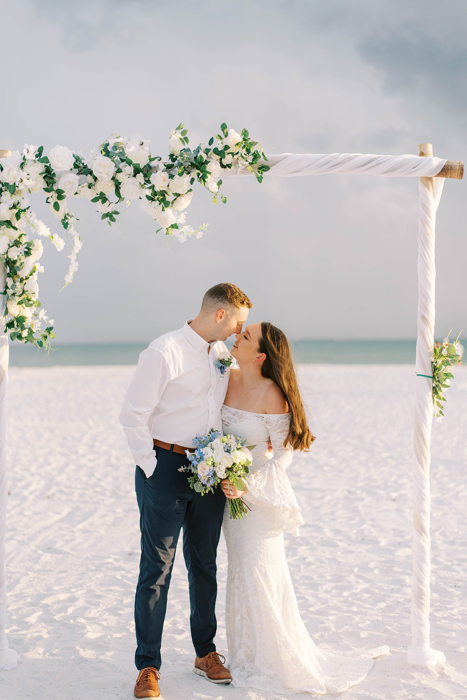 bride and groom kiss under floral arbor on beach wedding day