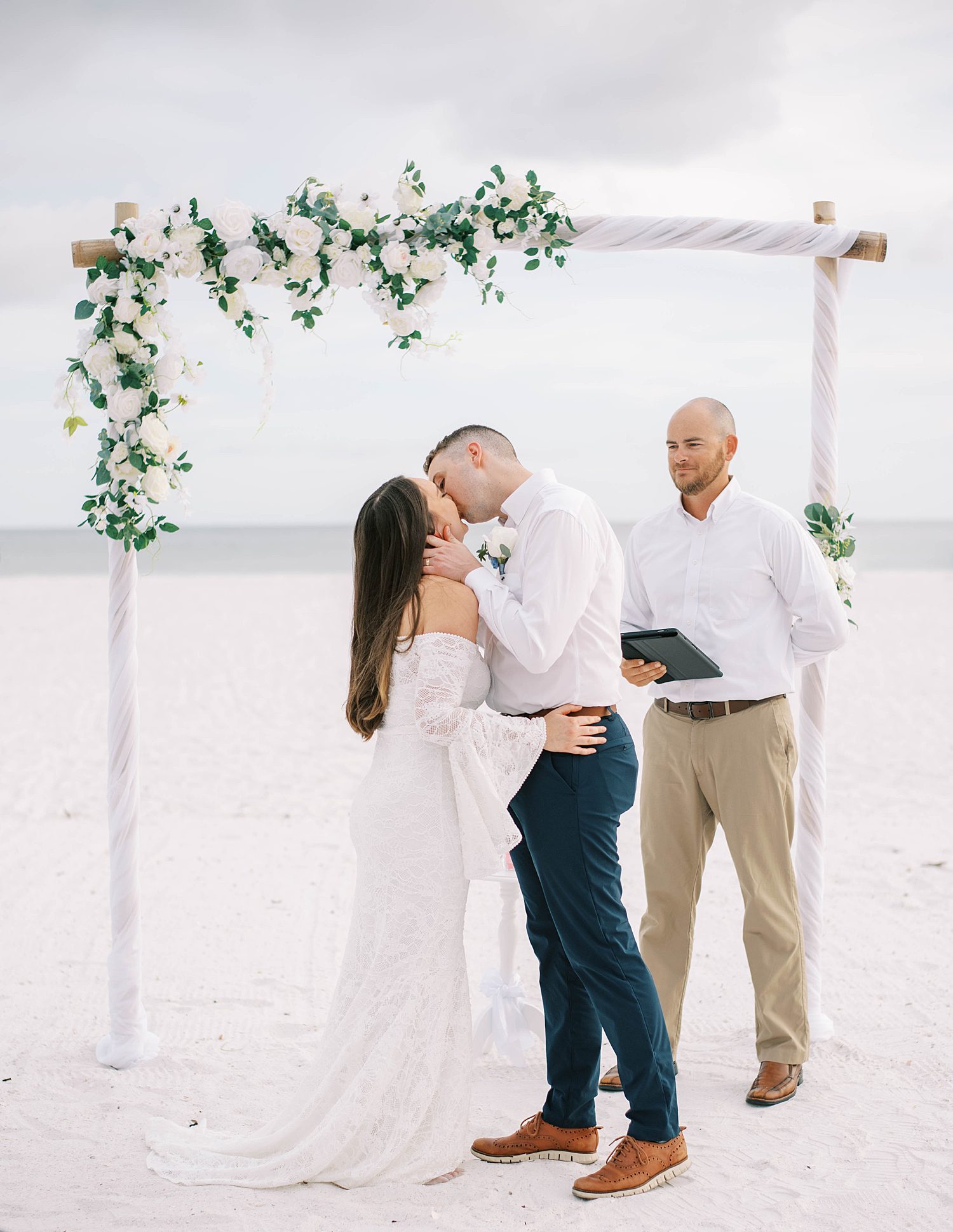 bride and groom kiss under floral arbor on beach
