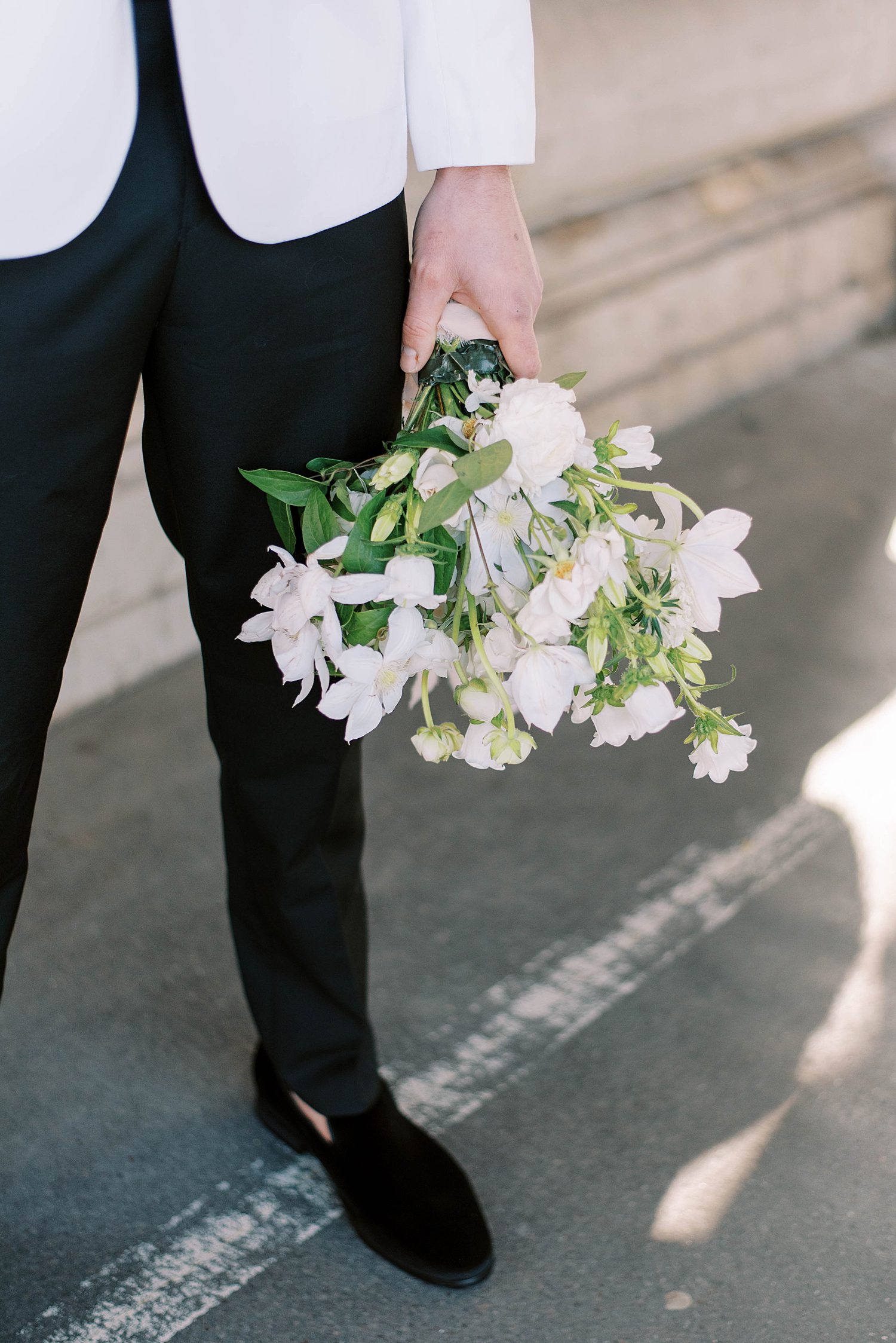 groom in white tux jacket holds white bouquet of flowers in Paris