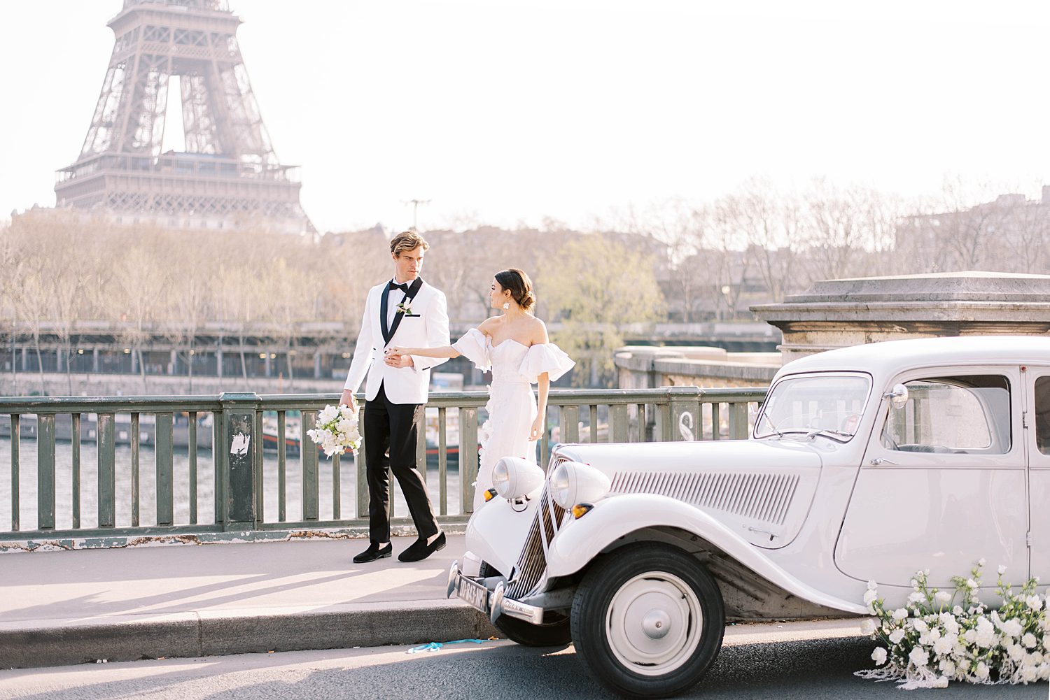 groom leads bride along bridge in Paris by white classic car