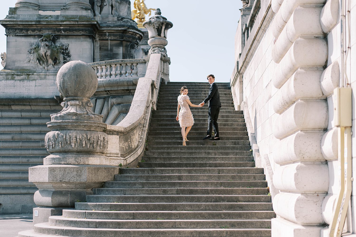 bride and groom hold hands walking up steps in Paris in black tux and blush Marchesa gown