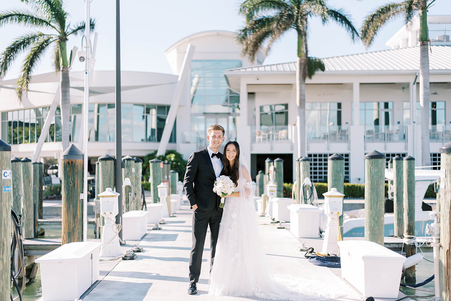 bride and groom stand on dock between palm trees outside Sarasota Yacht Club