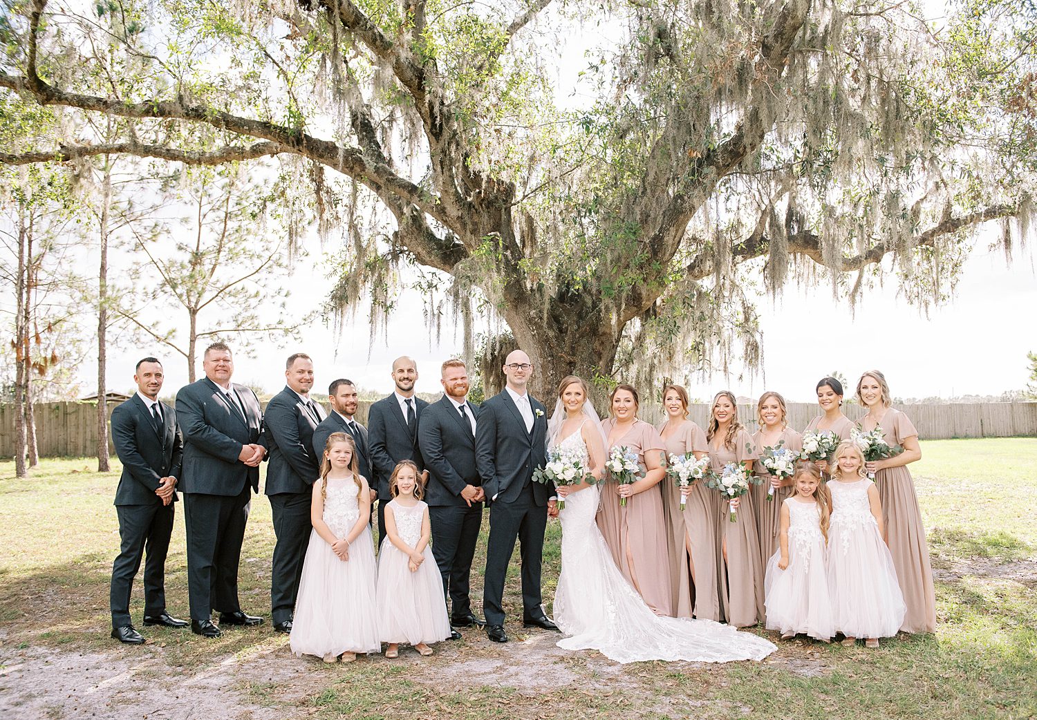 bride and groom stand by oak tree with wedding party in champagne and grey suits