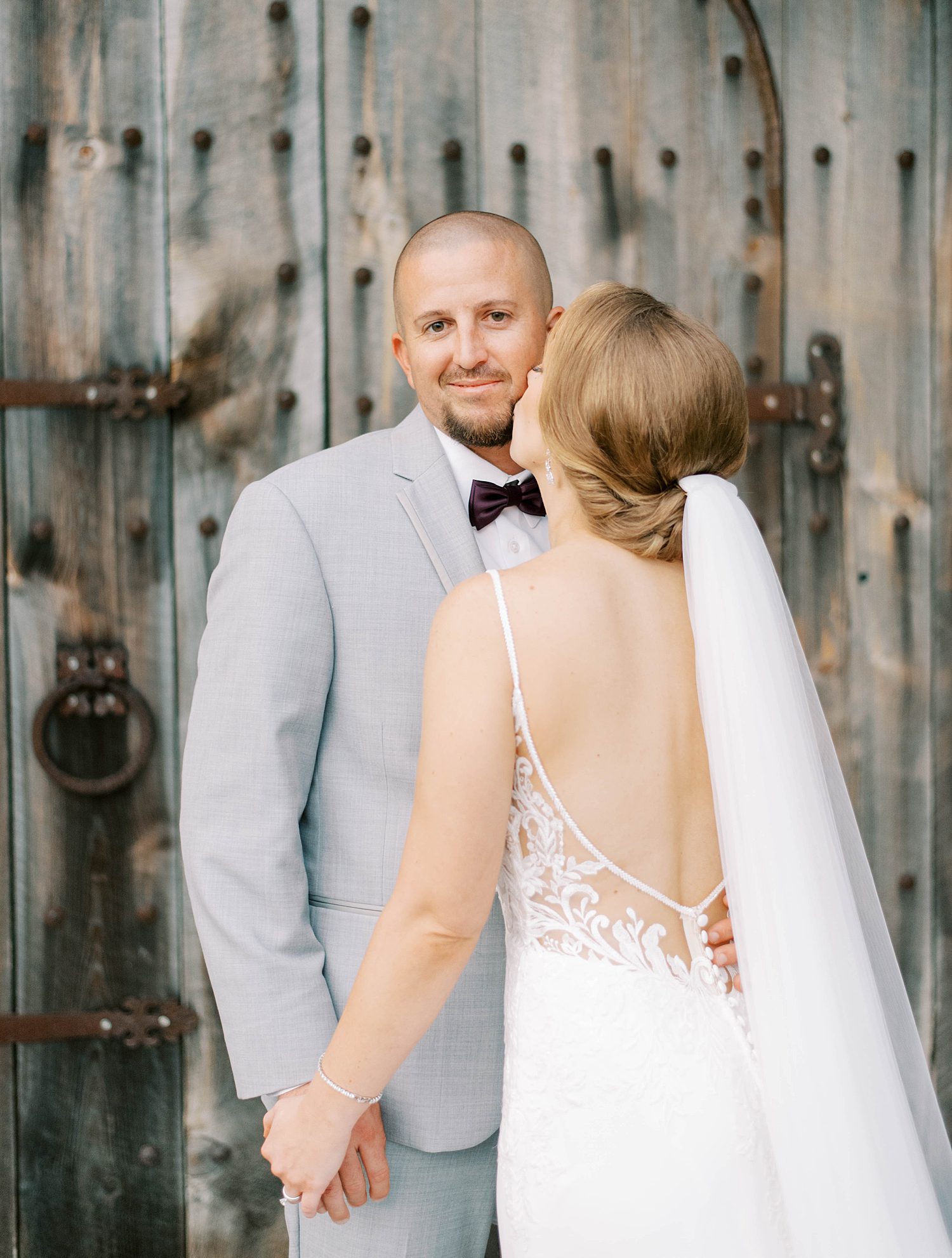 bride leans up to kiss groom's chin by wooden door at Bella Cosa Lakeside