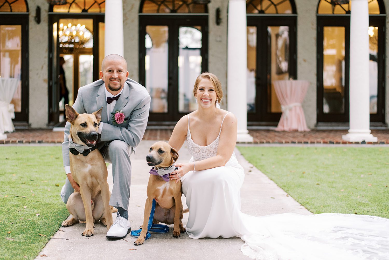 bride and groom kneel with dogs in front of Bella Cosa Lakeside