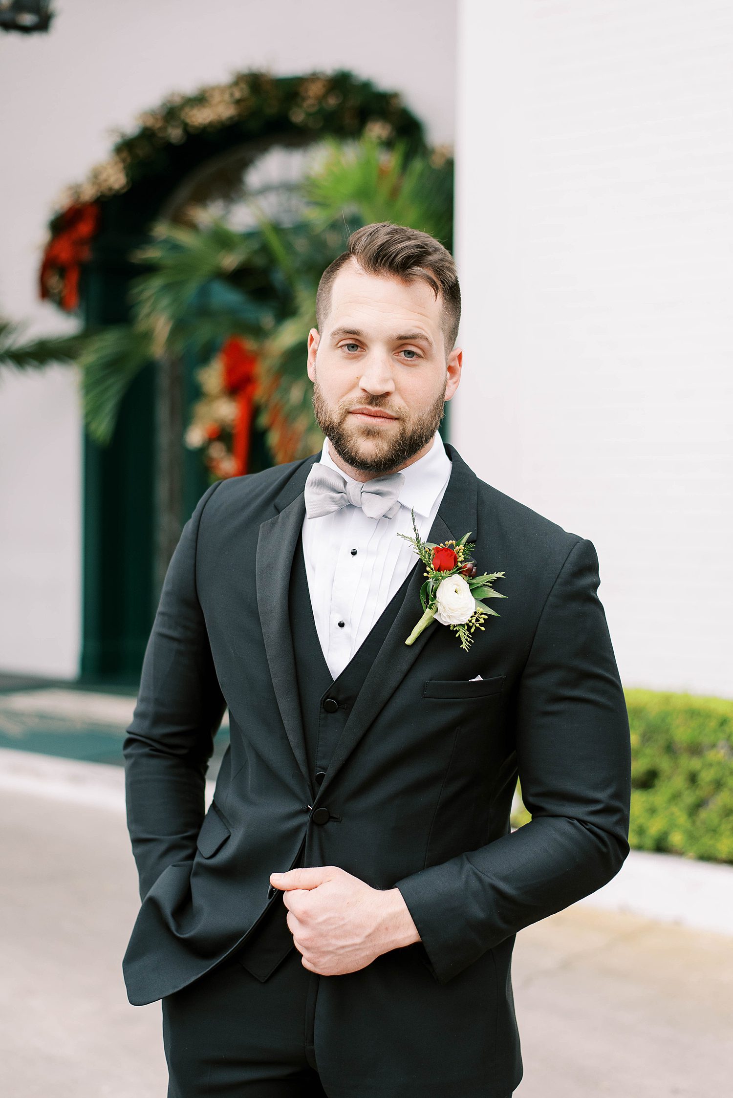 groom poses in black suit with red and white flowers 