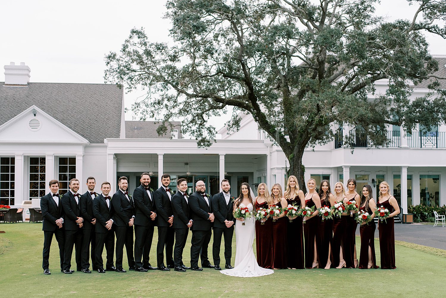 bride and groom stand with wedding party in red velvet gowns and black tuxes outside Palma Ceia Country Club