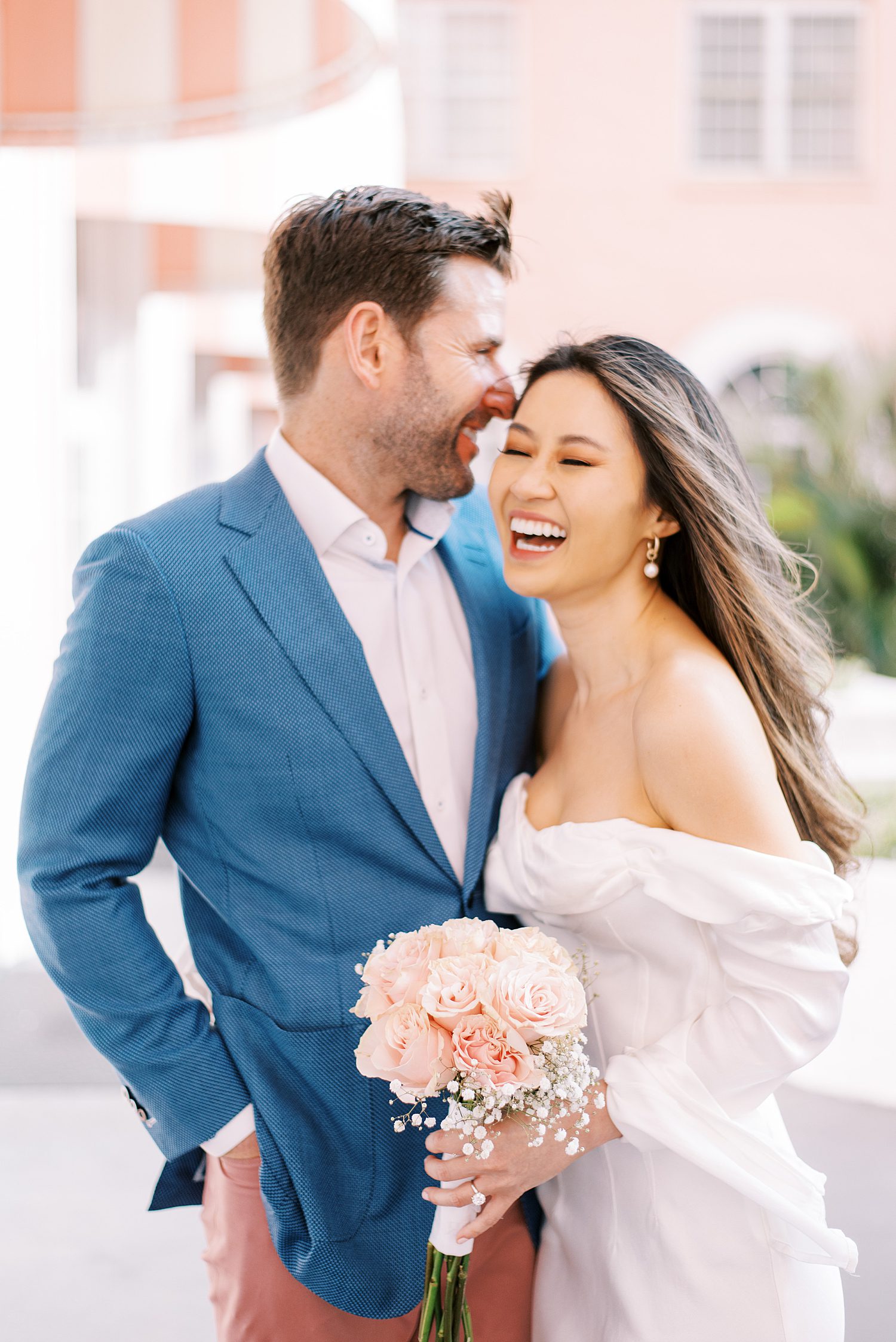 man in blue suit jacket makes woman in off-the-shoulder white gown laugh during St. Pete Beach engagement session