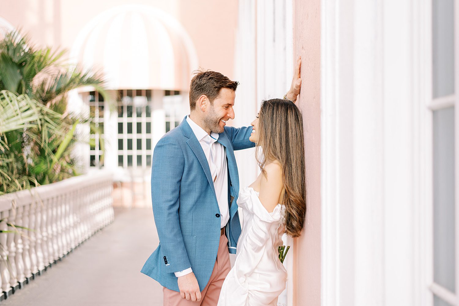 woman in white dress leans against pink wall at the Don CeSar Hotel with man in blue suit jacket leaning over her