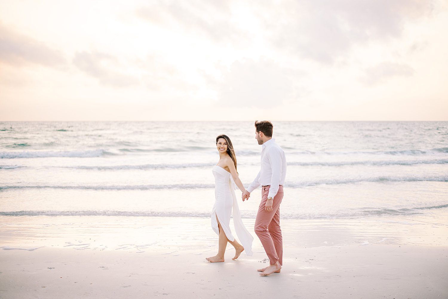 woman in strapless white gown walks on beach in Florida at sunset leading fiancé behind her 