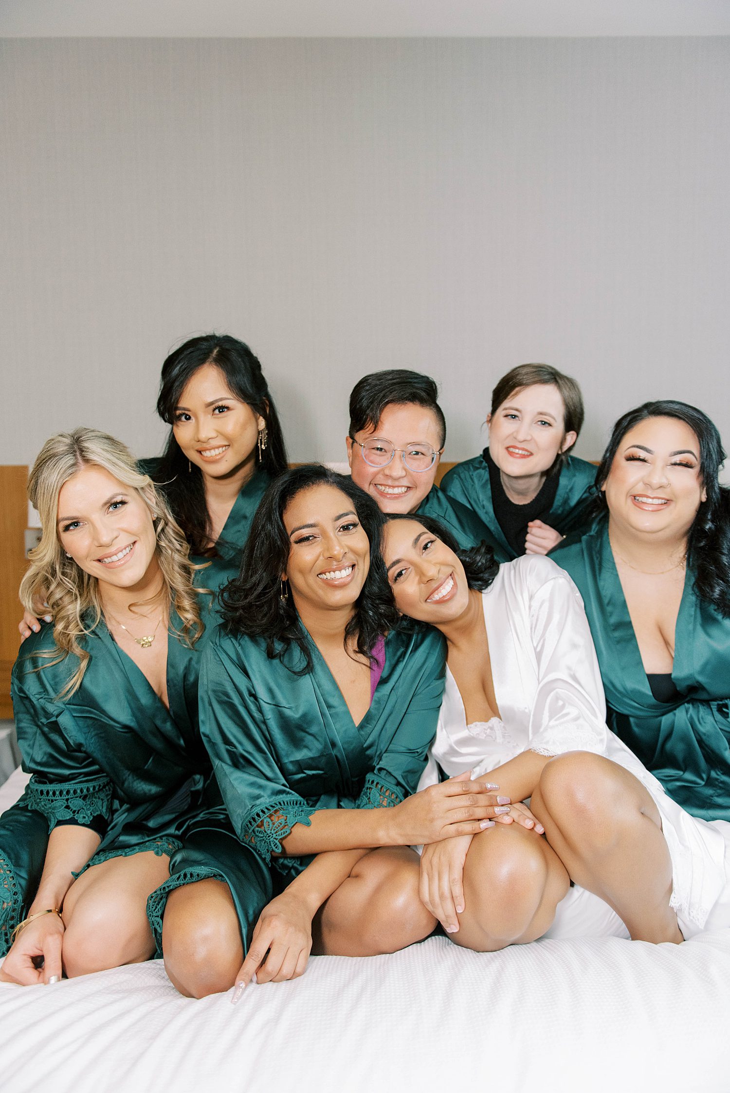 bride smiles with bridesmaids in teal robes leaning on maid of honor's shoulder