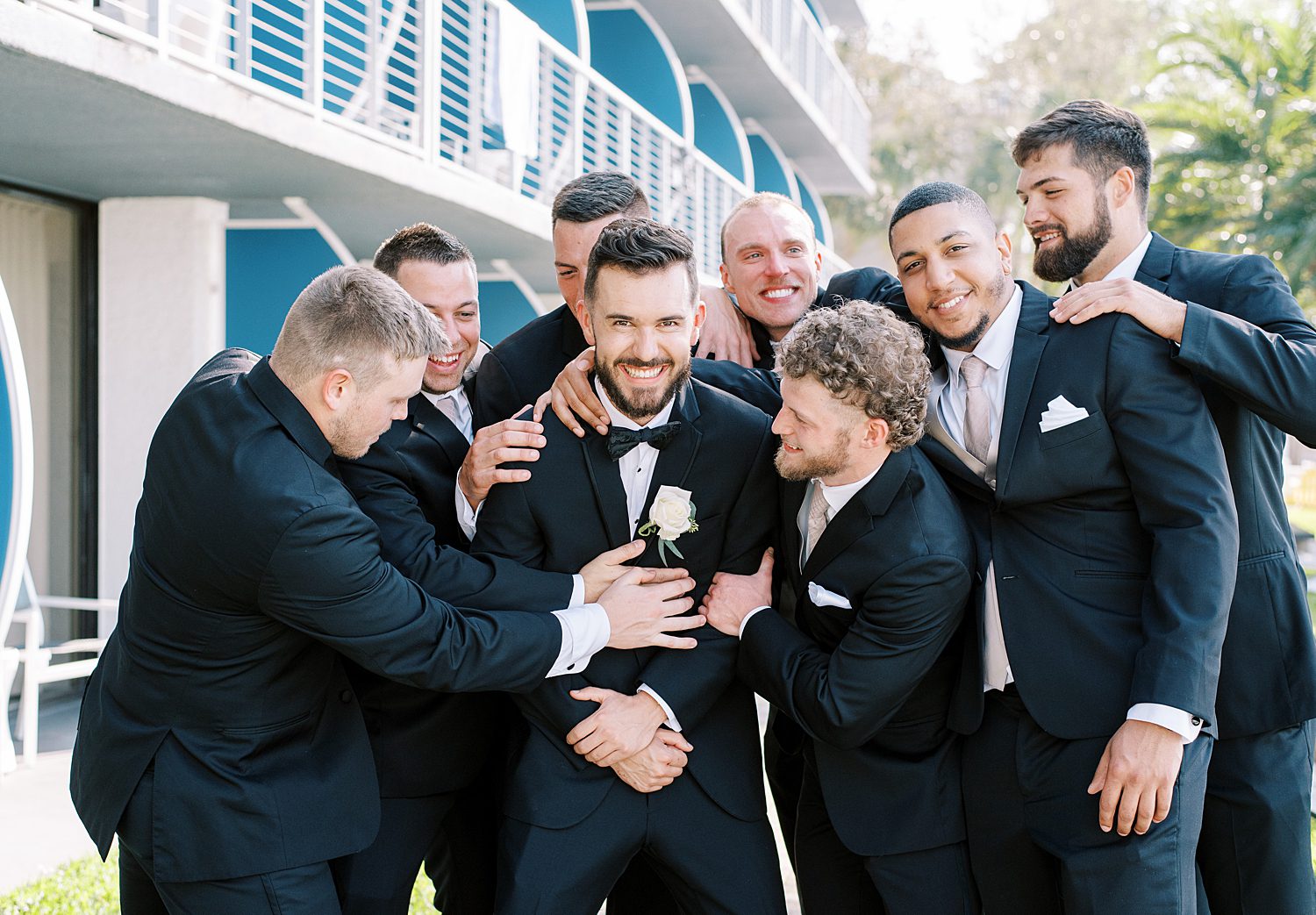 groomsmen hug and laugh with groom in black suits