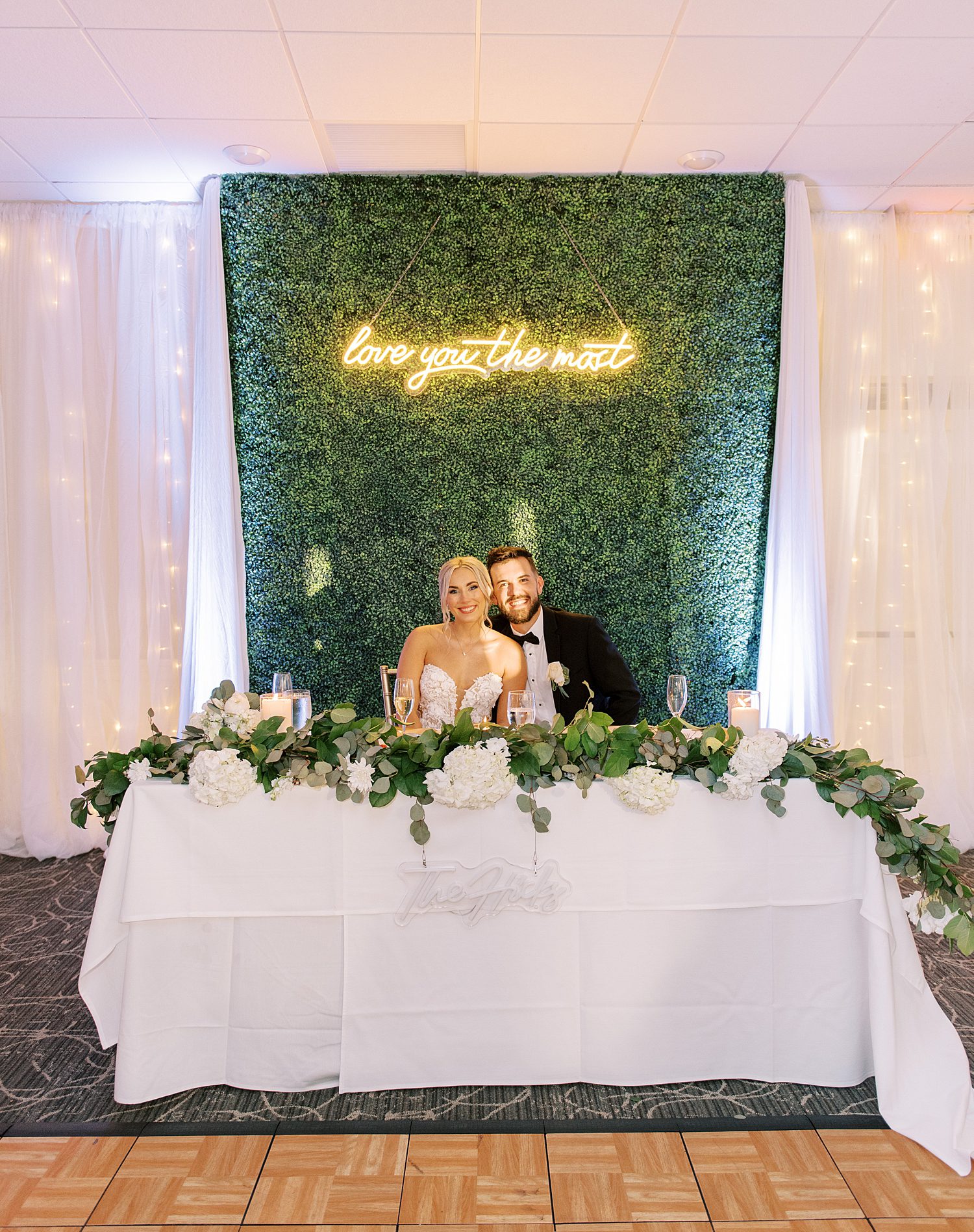 bride and groom sit in front of greenery wall with neon sign during Tampa FL wedding