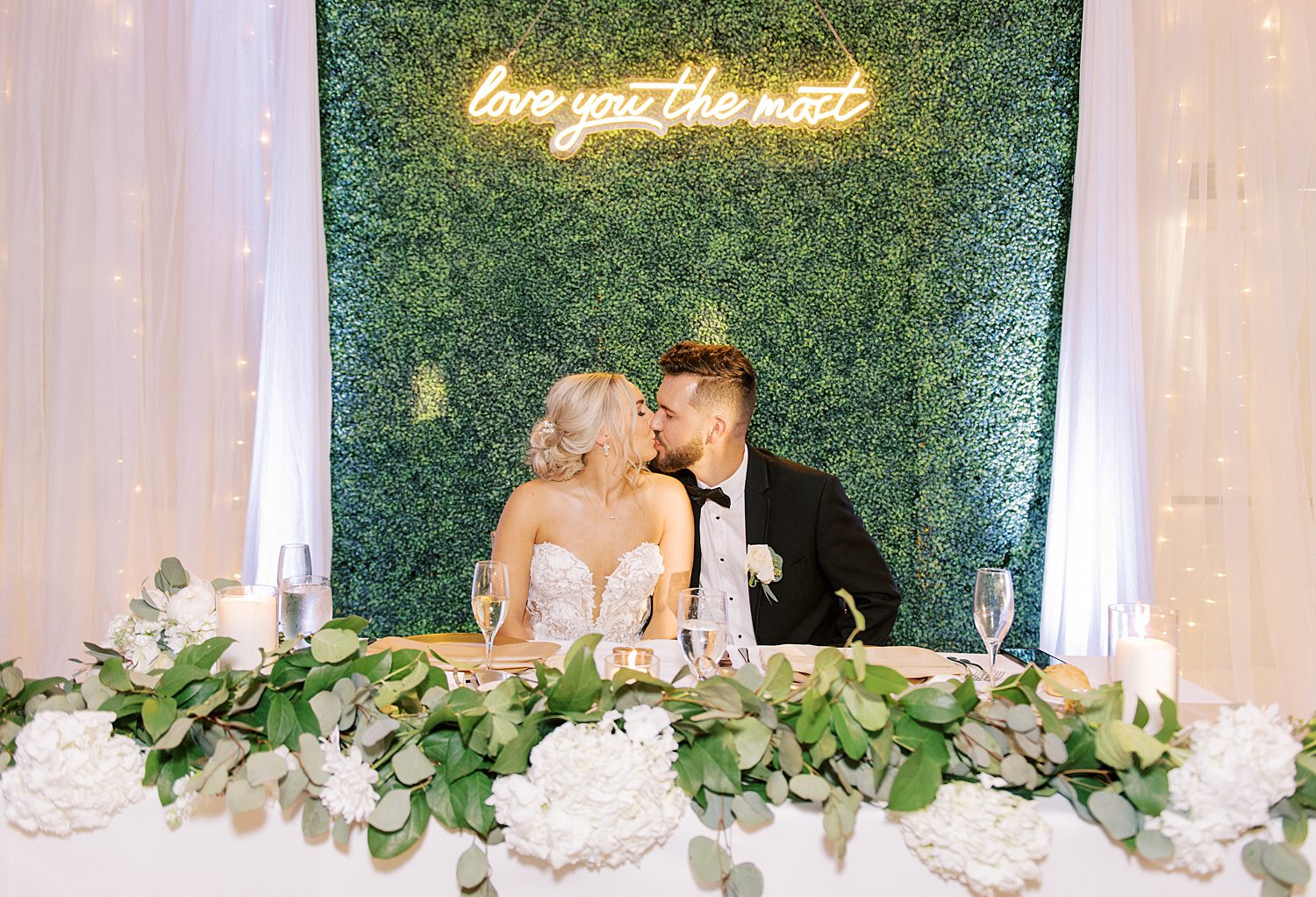 bride and groom kiss by greenery wall with neon sign