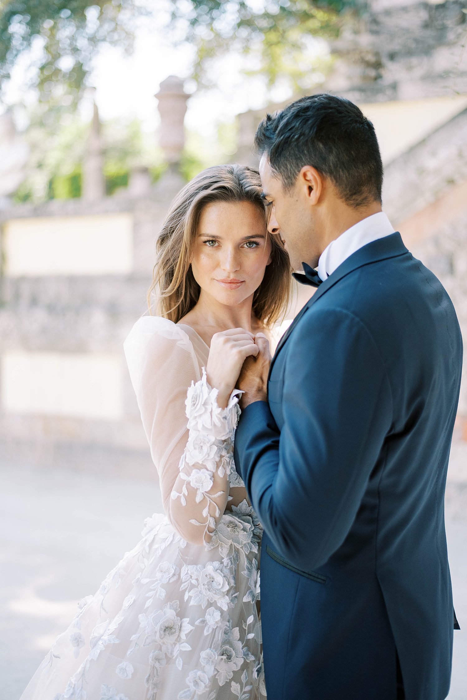 groom in navy suit hugs bride with lace sleeves at Vizcaya Museum