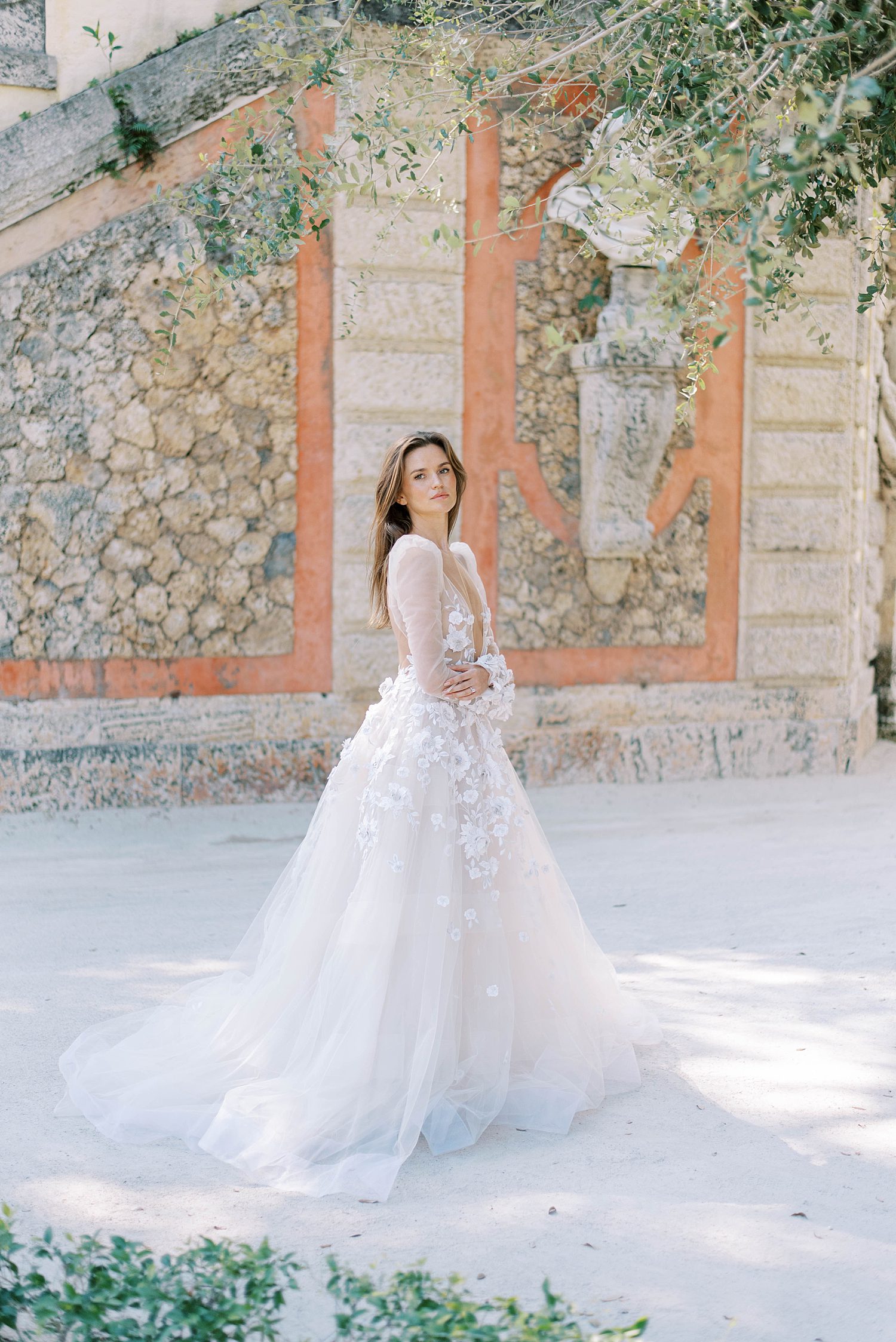bride stands in front of stone wall with long-sleeve lace wedding dress