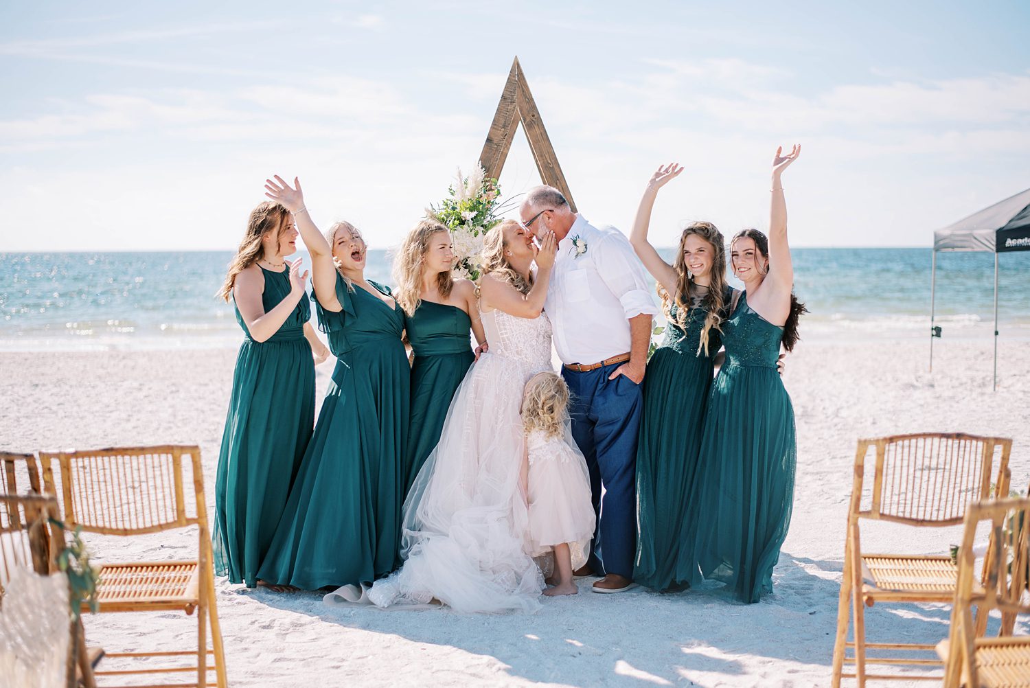 newlyweds kiss by wooden arbor with daughters in teal dresses cheering