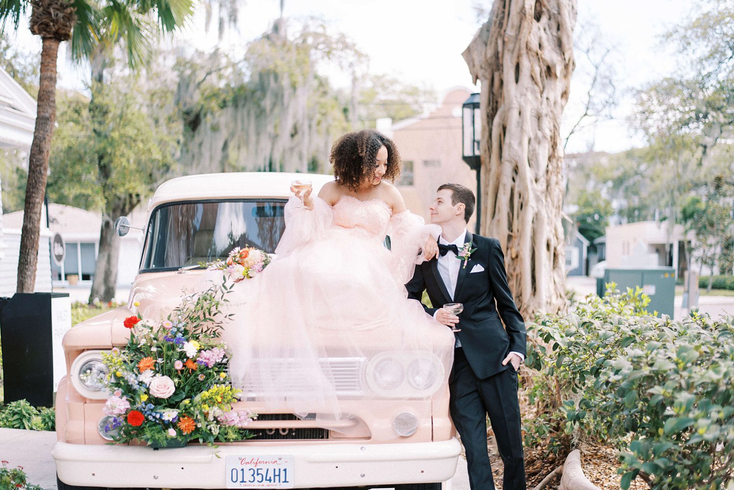 groom in black tux looks up at bride sitting on vintage pink truck