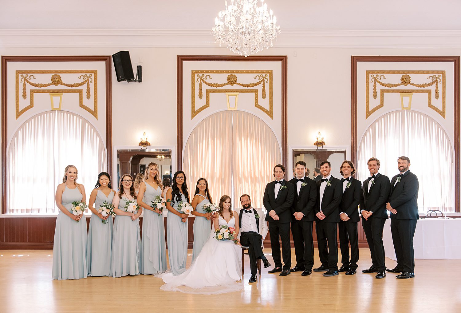 newlyweds sit between wedding party in light green and black suits