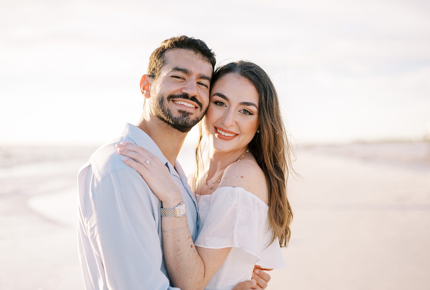 engaged couple hugs leaning heads together standing on beach at sunset
