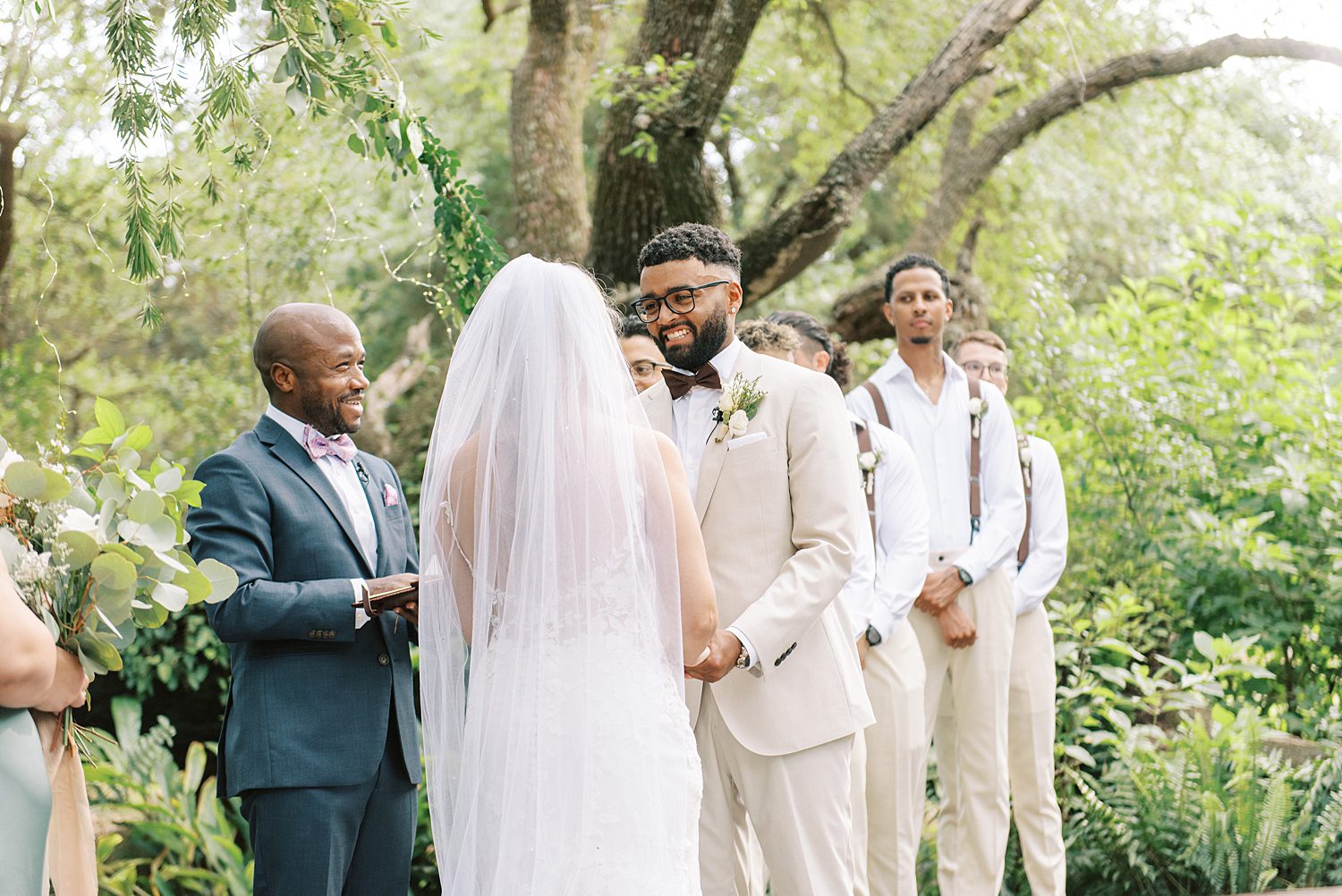 groom in tan suit smiles at bride during wedding ceremony under oak tree at Mill Pond Estate