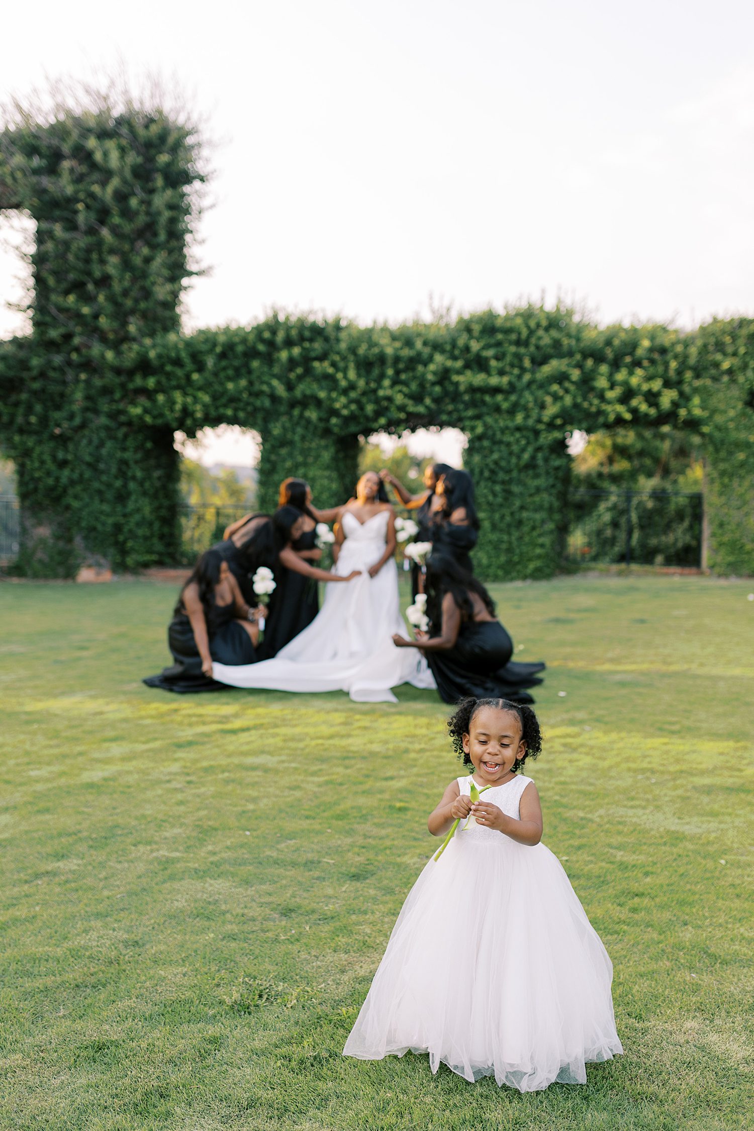 flower girl in white dress walks in front of bride and bridesmaids in black gowns