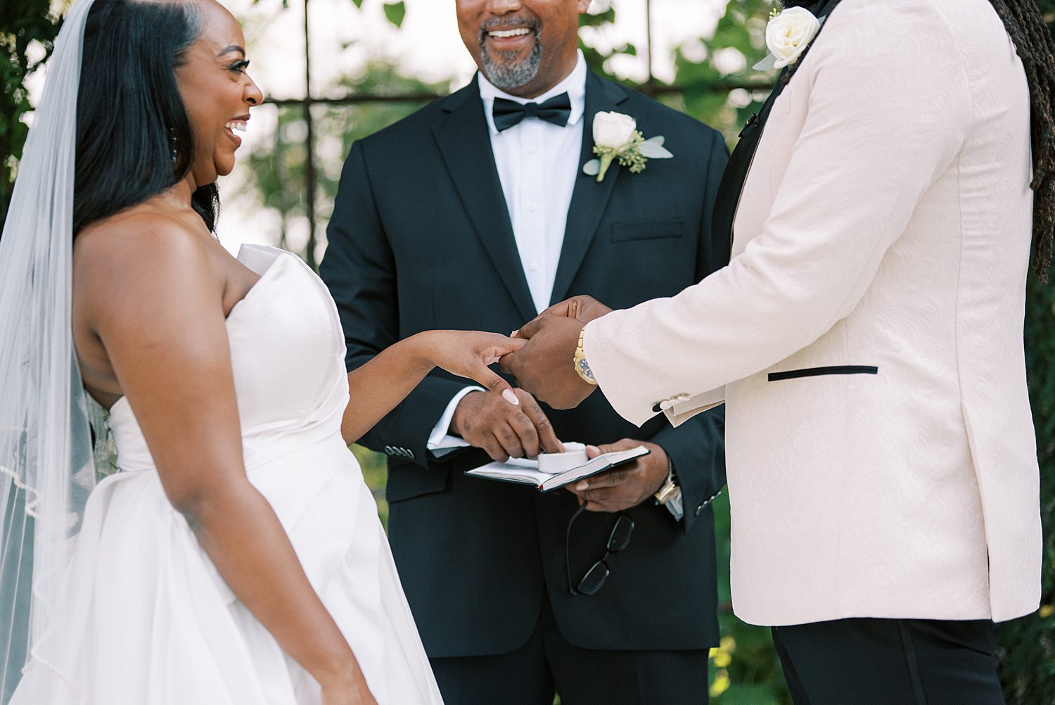 groom in white suit jacket puts ring on bride's finger during ceremony at The Bibb Mill Event Center wedding 