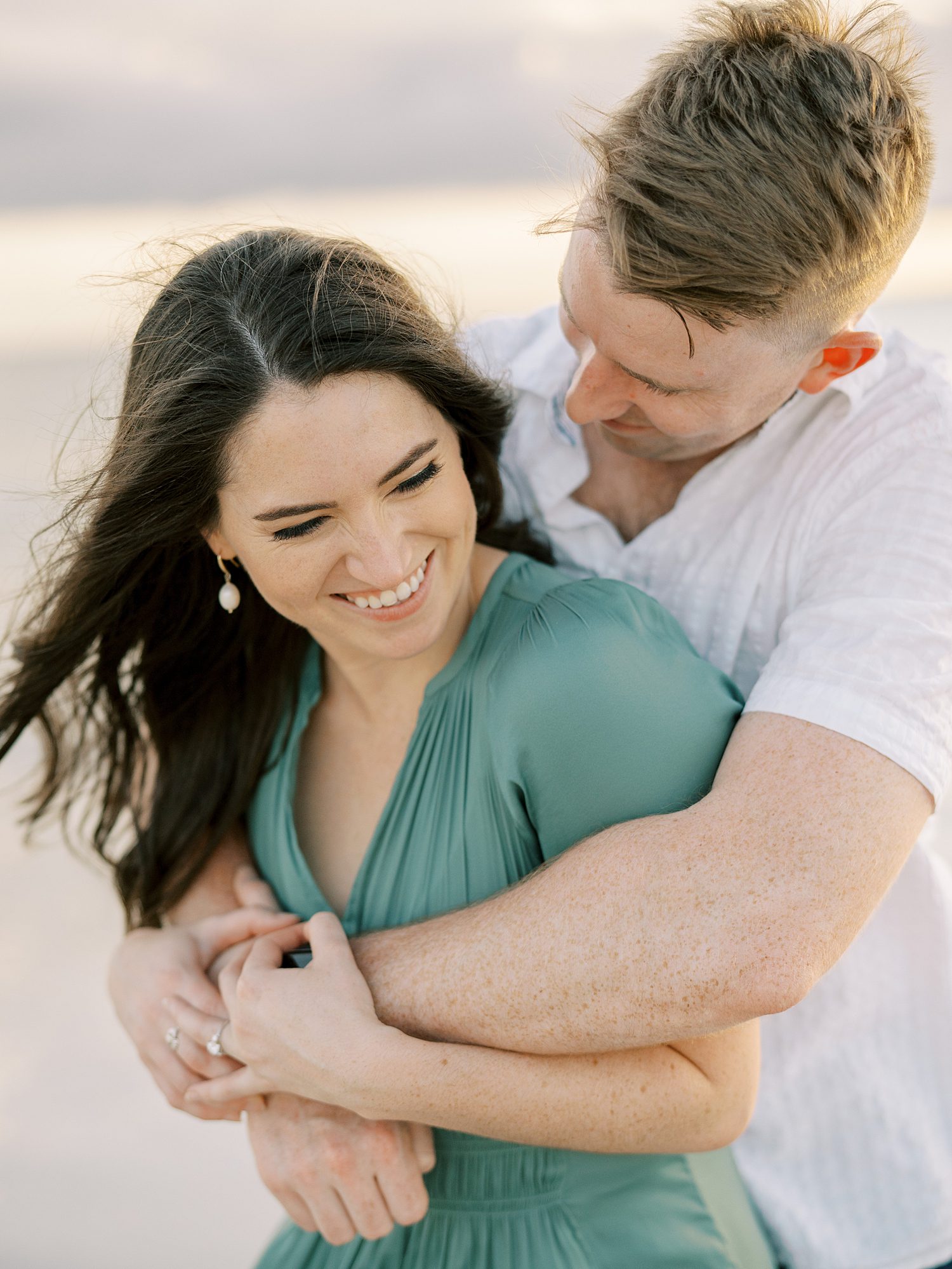 man in white t-shirt hugs fiancee on beach 