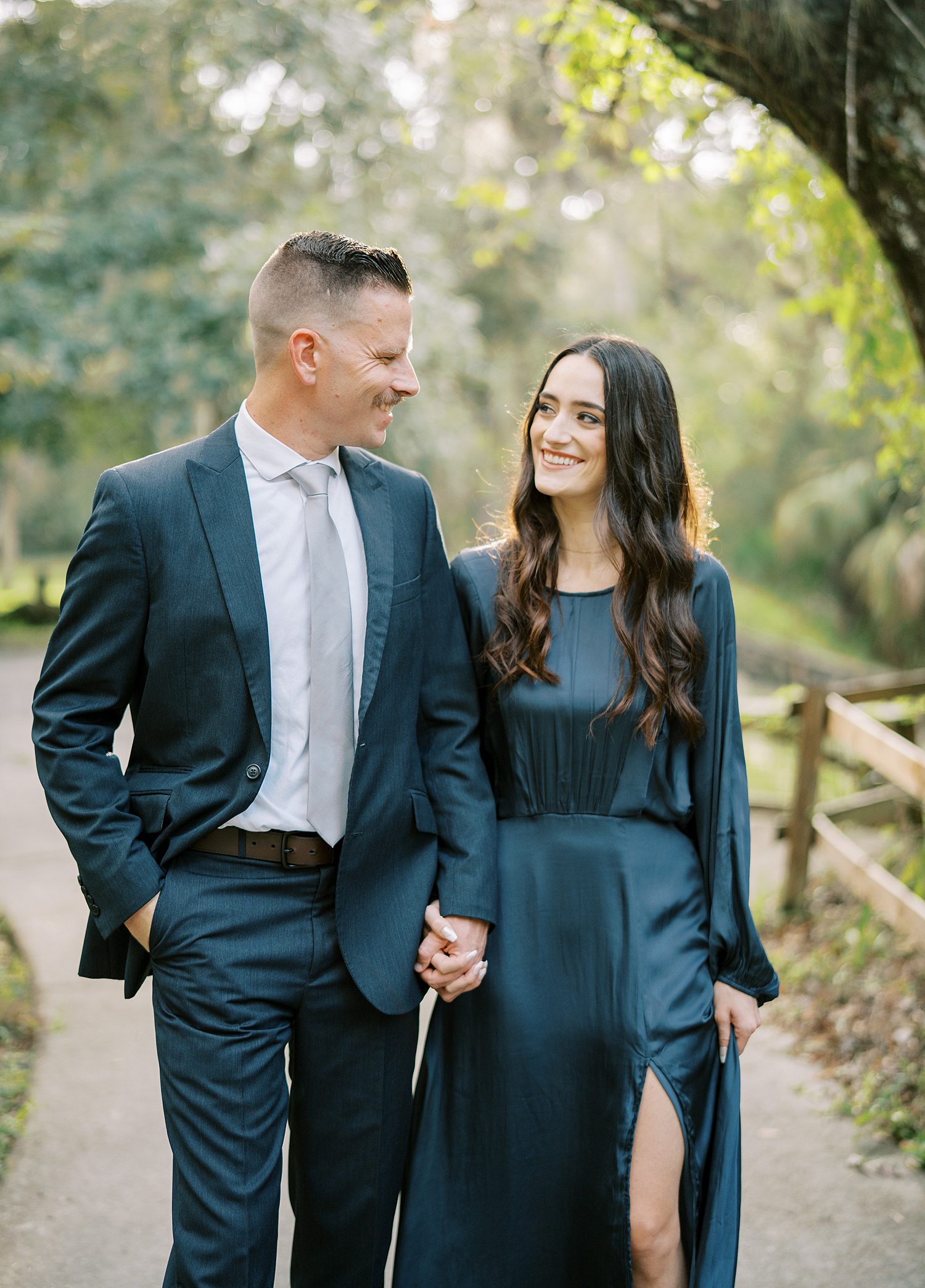 engaged couple holds hands in teal outfits walking through Hillsborough River State Park