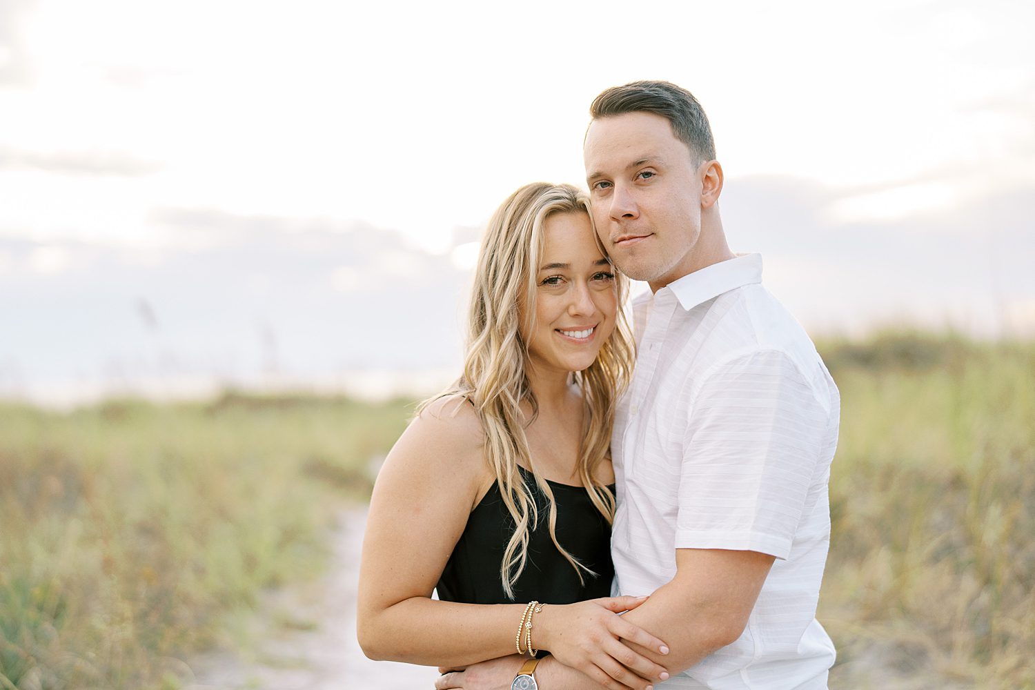 man hugs woman in black dress on sand dune during beach engagement session in Tampa FL