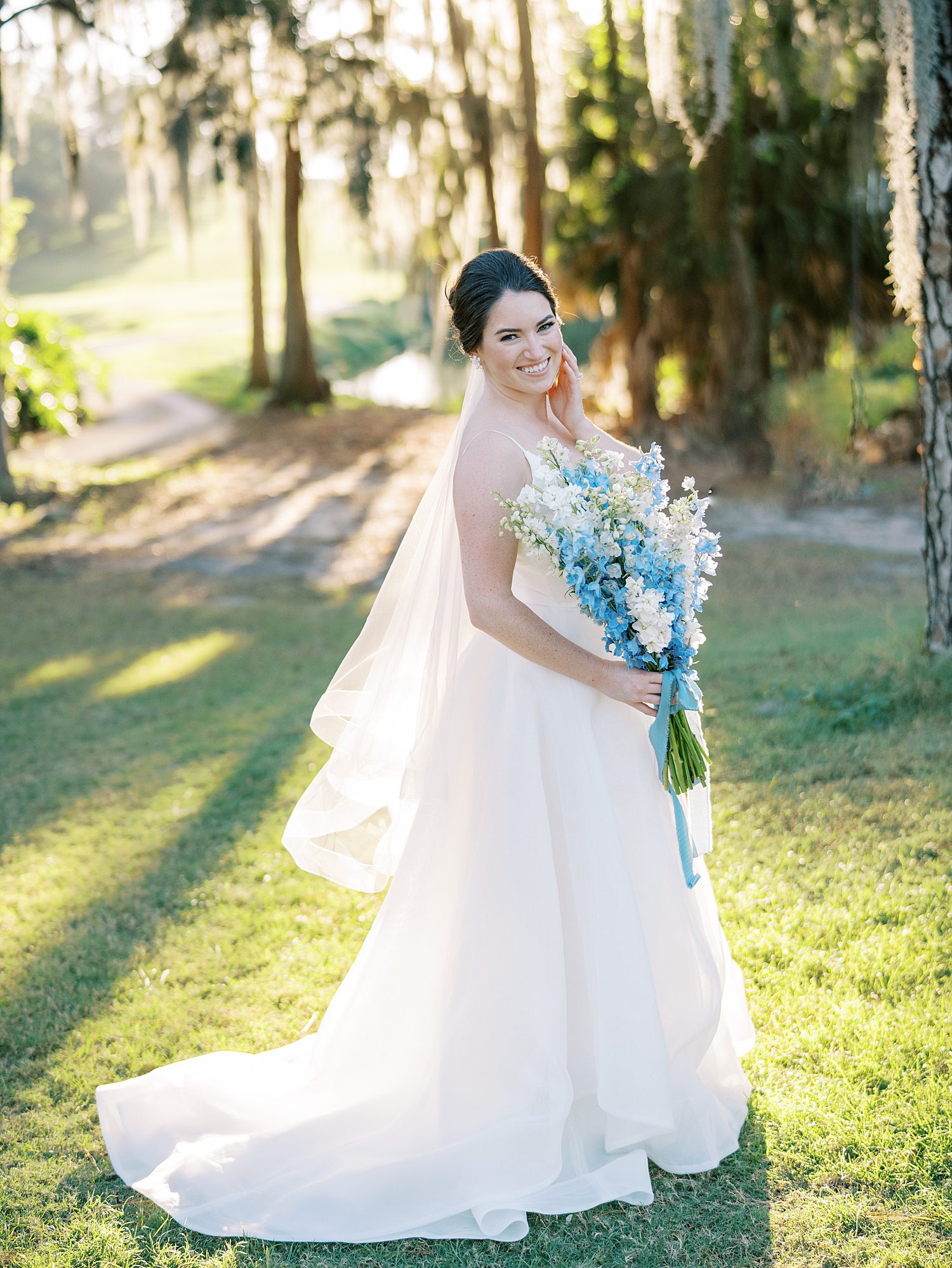 bride smiles looking over shoulder holding long blue bouquet of summer flowers 