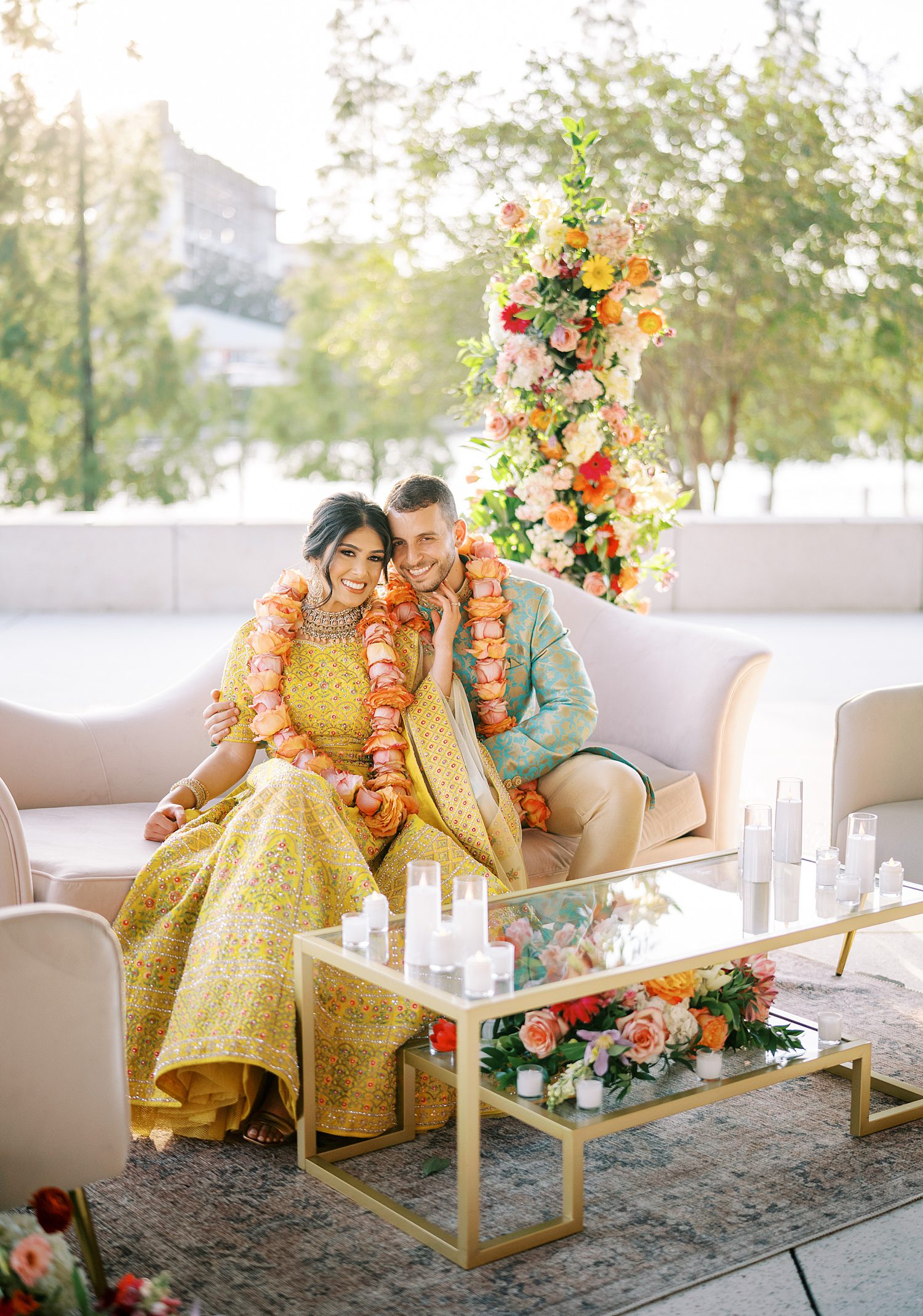 newlyweds sit on white couch in front of colorful flower arrangements during Mehndi Day at the Tampa Museum of Art