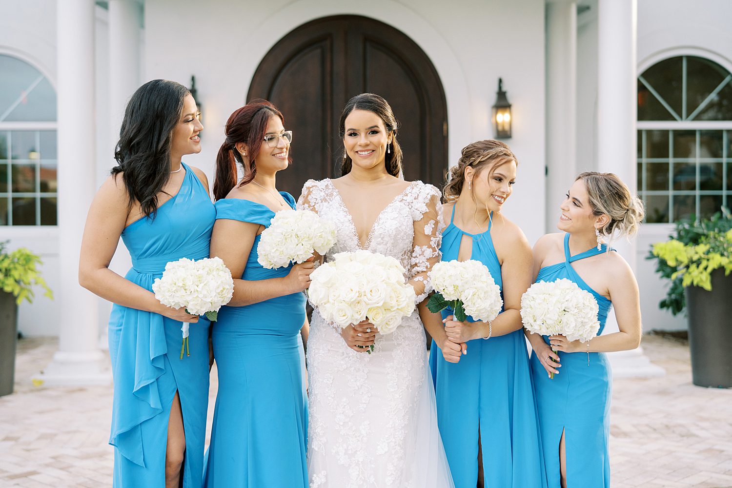 bride smiles with bridesmaids in blue gowns with white flowers outside Harborside Chapel