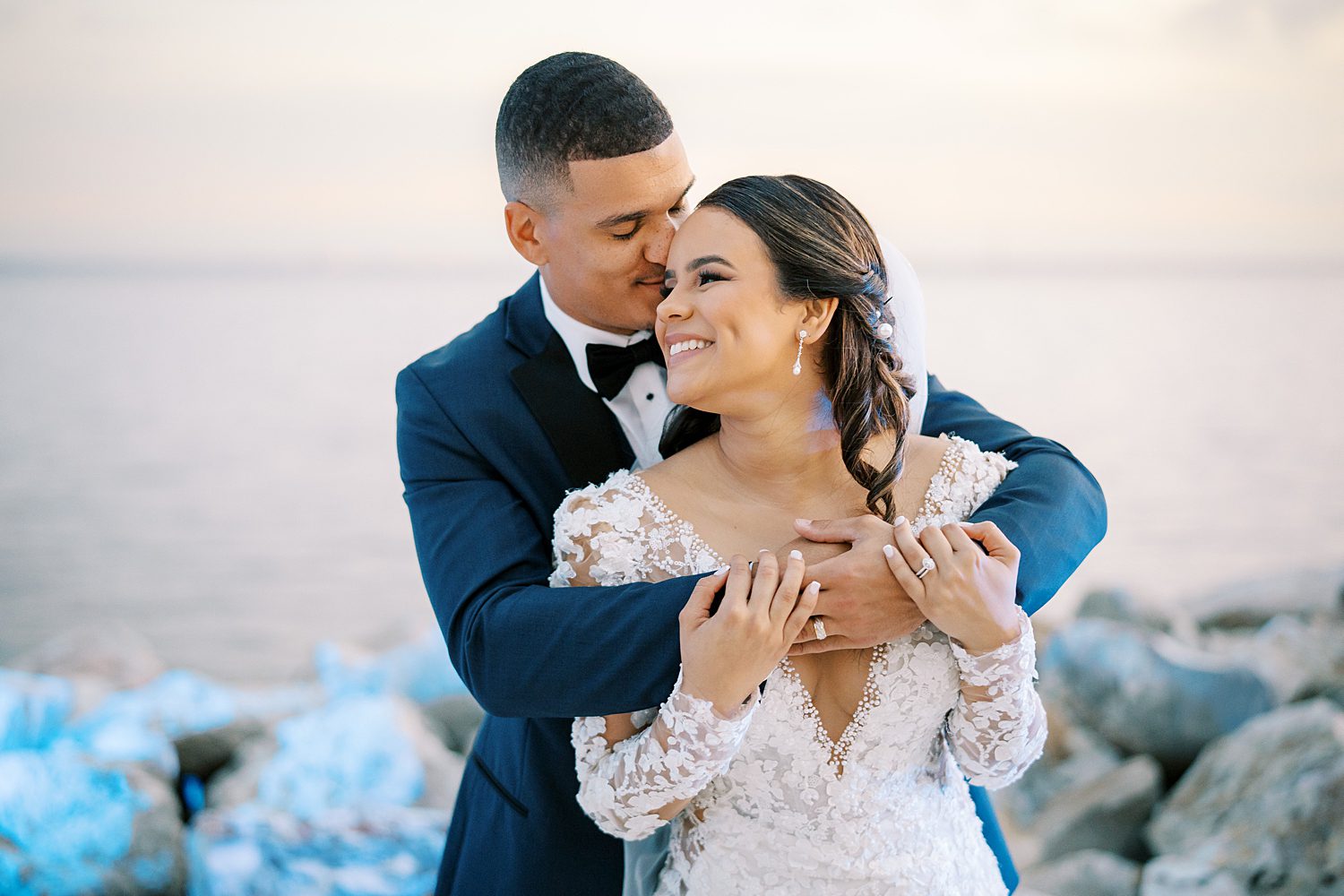 groom hugs bride from behind in front of rocks outside the Rusty Pelican