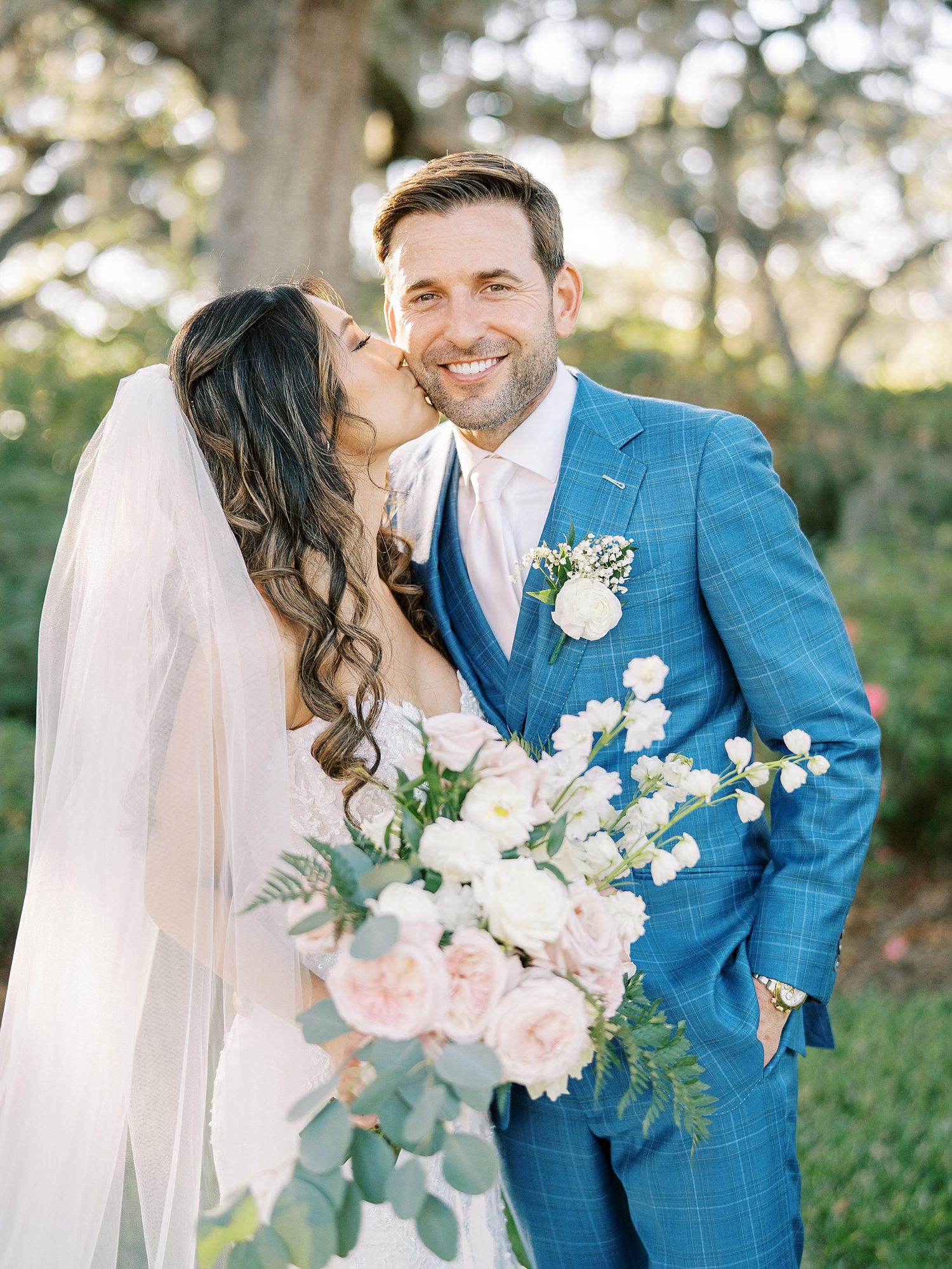 bride leans up to kiss grooms cheek while holding blush and ivory flowers 