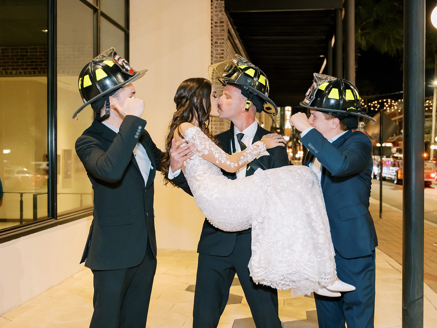 bride and groom kiss with firemen buddies around them 