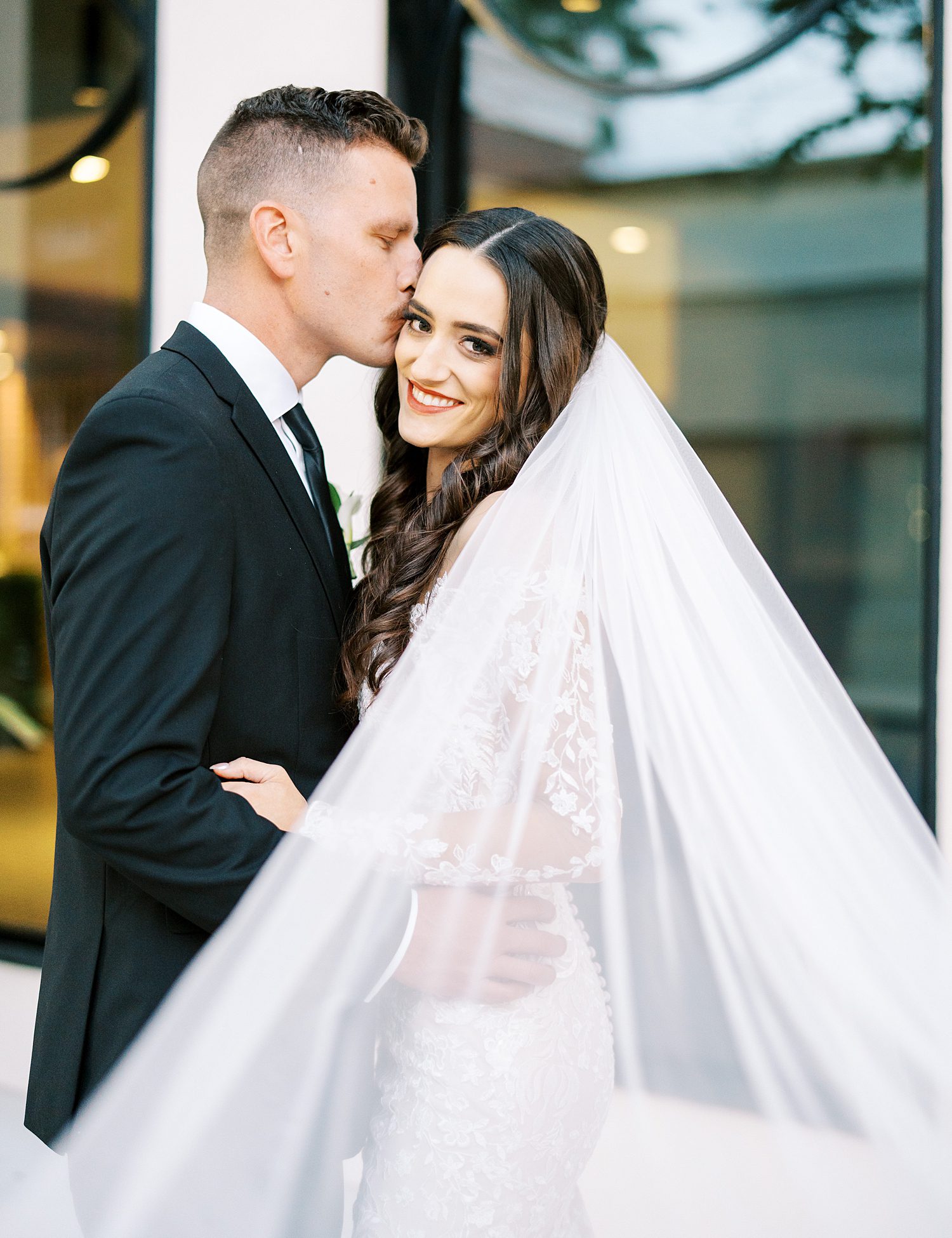 groom kisses bride's cheek with veil pulled around them in front of the Hotel Haya