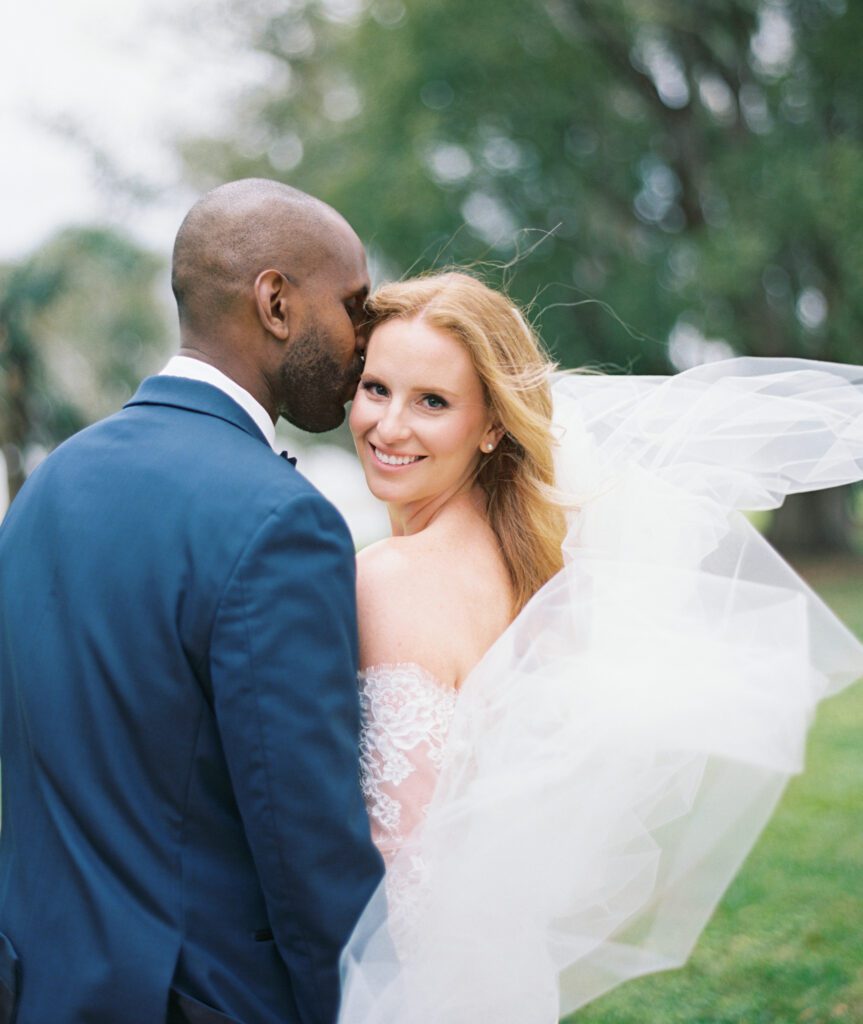 Adams Estate Wedding Photographer film photography in Tampa Florida Bride looking at camera with wind lifting up her veil, Groom kissing her temple