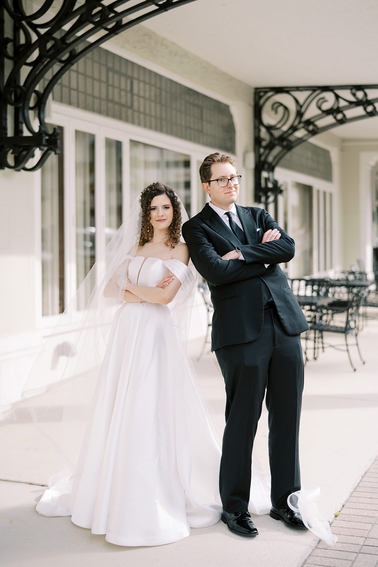 newlyweds stand leaning back to back with arms crossed