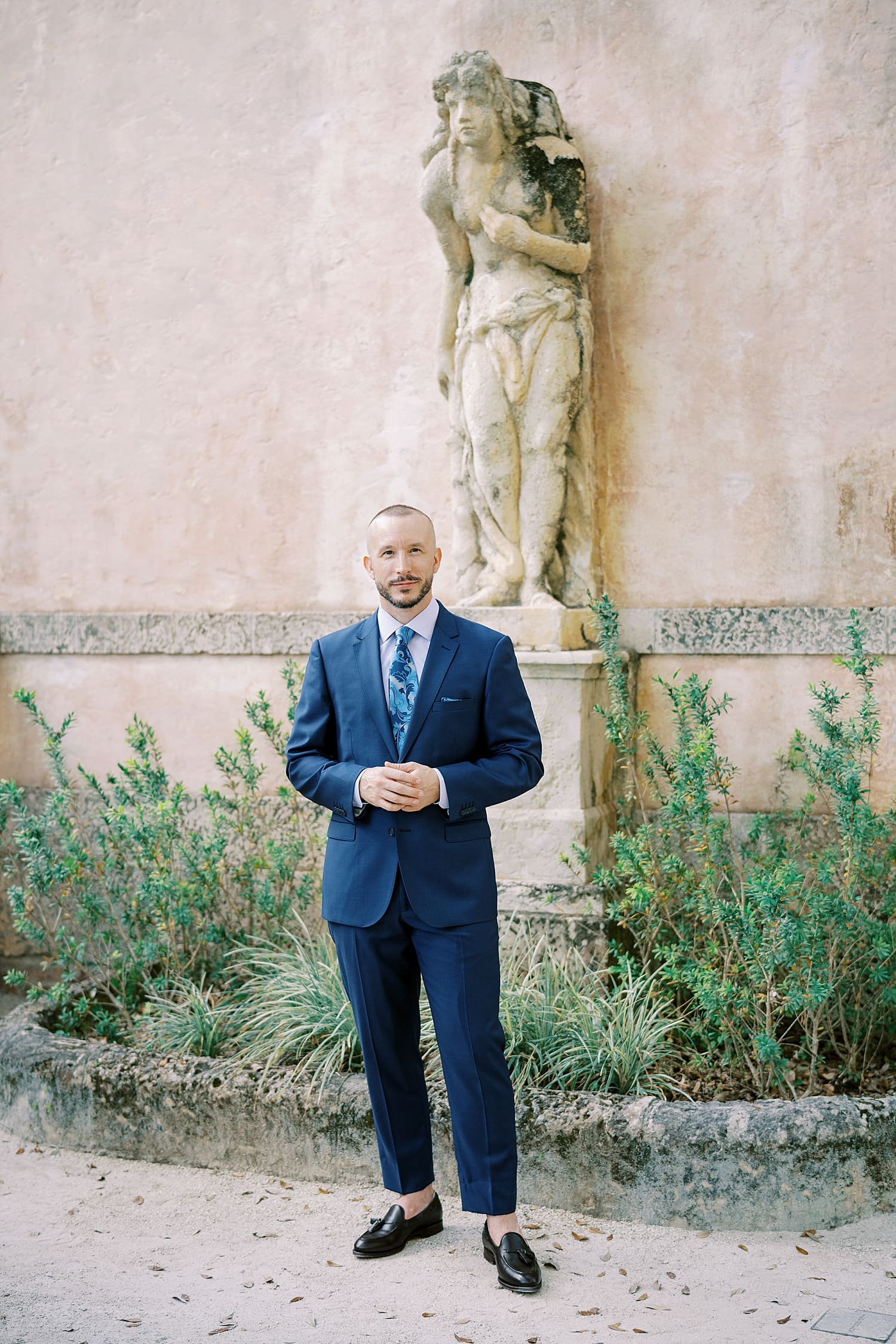 man in navy suit stands by statue at Vizcaya Museum  