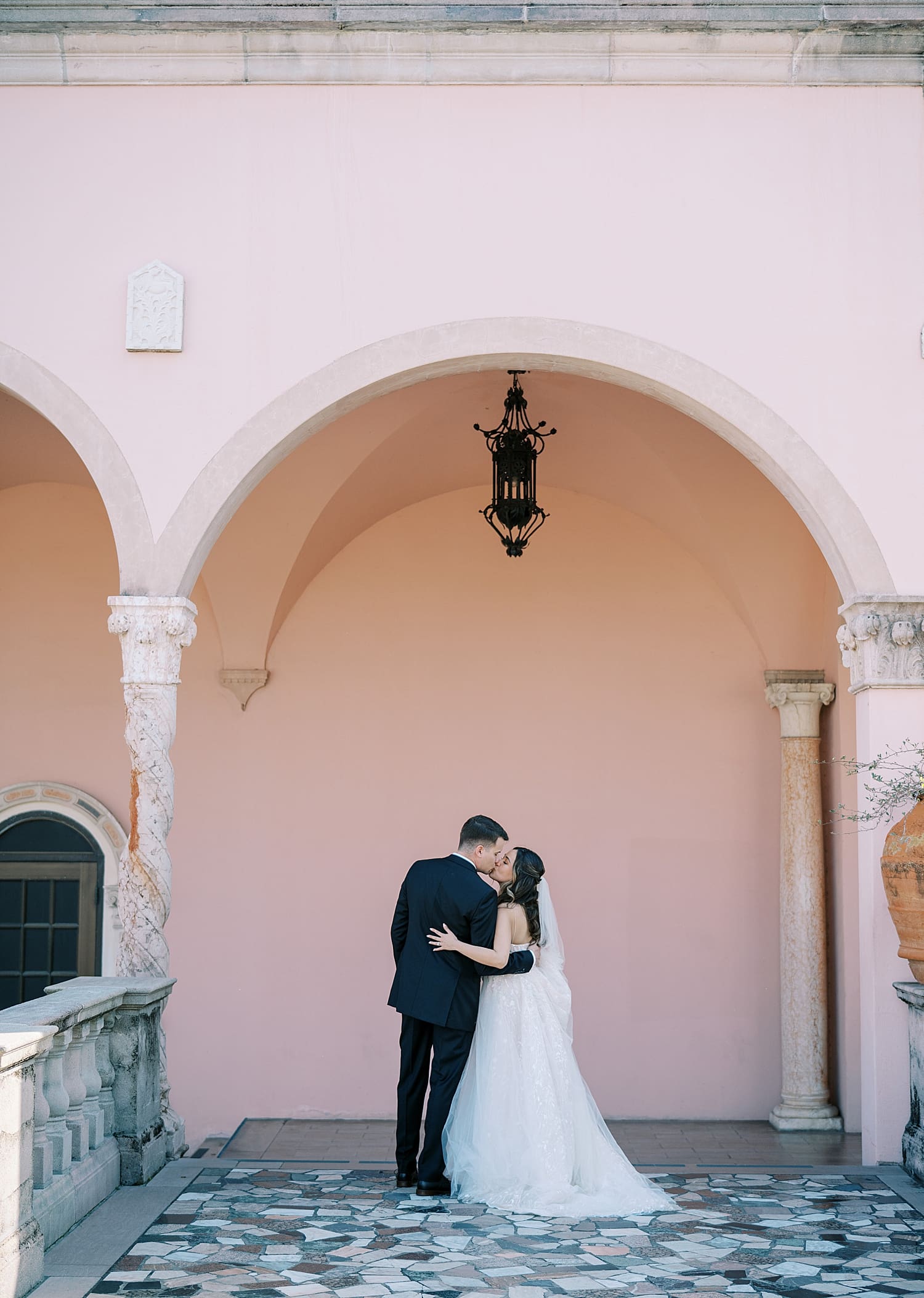 newlyweds kiss under archway of pink exterior building of The Ringling Museum
