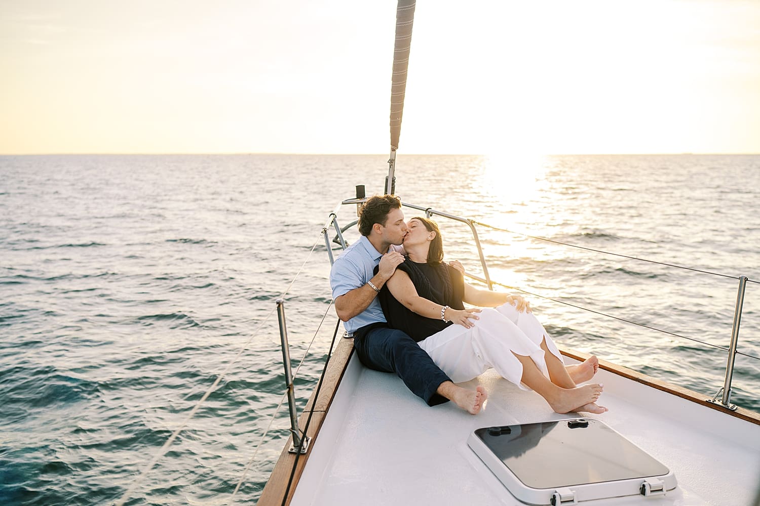 couple kiss while sitting on front of sailboat as the sun rises behind them