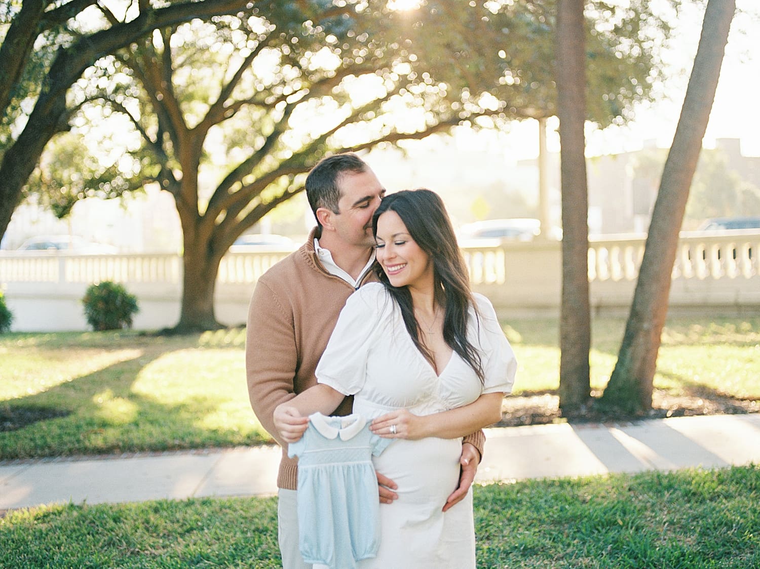 husband kisses his wife's head as she holds up blue outfit for baby boy