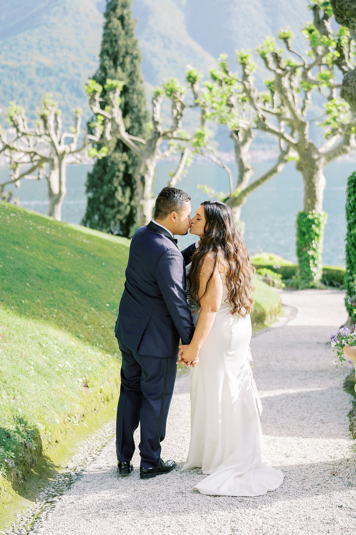 newlyweds kiss on path by Lake Como lined by trees and greenery
